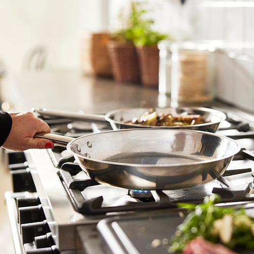 Una mano sostiene una sartén de acero inoxidable en una estufa de gas. Otra sartén con alimentos para cocinar está detrás, probablemente usando la función de distribución de calor uniforme de las sartenes.