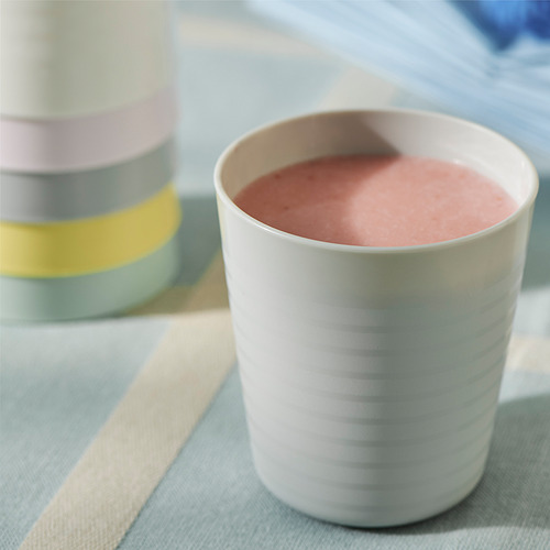 Una taza blanca para bebés se encuentra sobre una tela a rayas, que contiene batido rosa. Detrás de él, se ven coloridas tazas apilables.