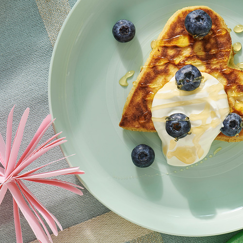 El plato verde claro para niños contiene un delicioso panqueque de arándanos cubierto con yogur y rociado con miel, todo colocado sobre un acogedor mantel azul.