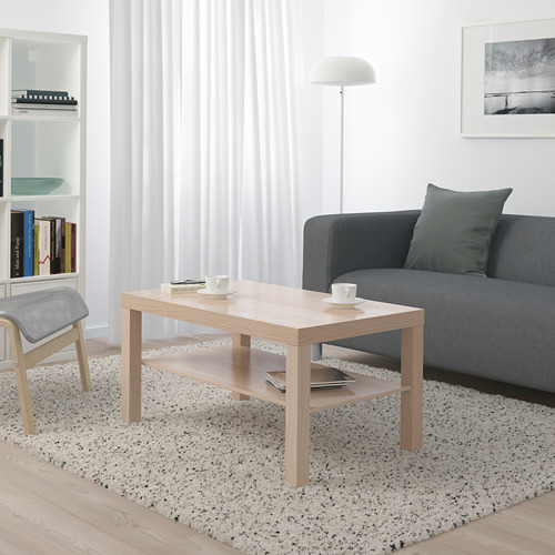 Salón minimalista: mesa de centro LACK, sofá gris con almohada verde, estantería blanca, lámpara de pie.