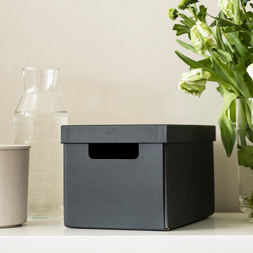 Una caja de tela gris con asa se encuentra sobre una mesa. Es ideal para organizar artículos pequeños como accesorios de escritorio.