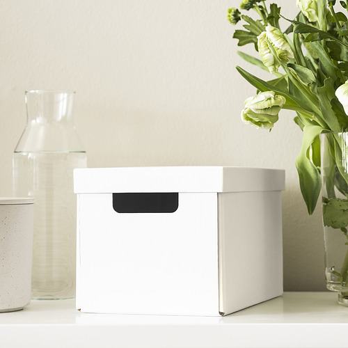 La caja blanca con asa negra se encuentra sobre una mesa blanca cerca de un jarrón de vidrio y flores verdes. Ideal para guardar objetos pequeños.