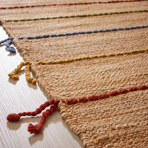 Alfombra de yute tejida a mano con coloridas rayas y borlas de lana, que muestra una intrincada artesanía y materiales de calidad.