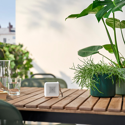 Pequeño altavoz blanco VAPPEBY se encuentra sobre una mesa de madera, rodeado de plantas y vasos para beber. Compacto y portátil.