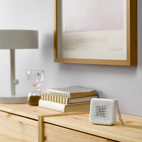 Pequeño altavoz blanco VAPPEBY se encuentra sobre una mesa de madera junto a libros y lámparas.