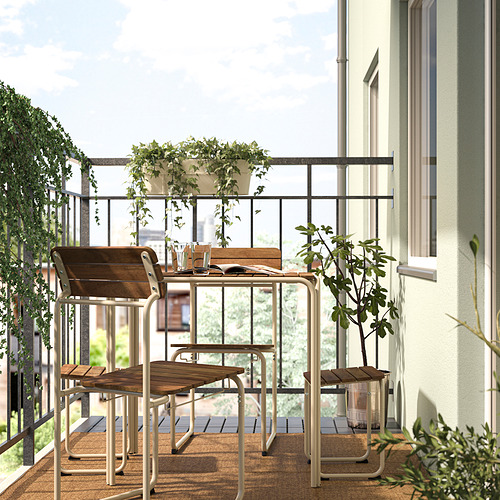 Balcón con plato de madera de acacia FURUÖN y metal blanco; Incluye mesa, sillas y reposapiés. Rodeado de plantas en macetas, creando un espacio acogedor.