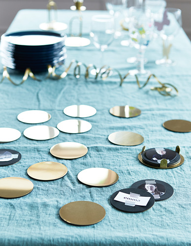 Mesa puesta con mantel azul, posavasos metálicos dispersos, algunos con nombres. Arriba a la izquierda, pila de posavasos azul marino en el soporte. Diseño elegante y reflectante.