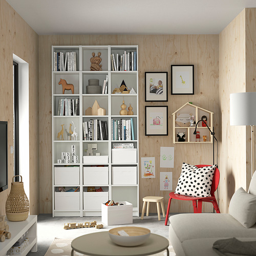 Sala de estar moderna con estanterías blancas BILLY, silla roja, almohada de lunares, paredes de madera y varios juguetes y libros para niños.