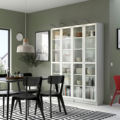 Comedor moderno con estanterías blancas con puerta de vidrio BILLY, sillas negras, paredes verdes, alfombra a rayas y lámpara colgante.