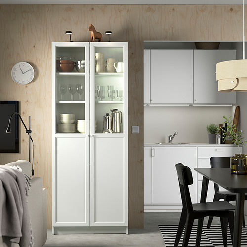 Cocina moderna con gabinete BILLY blanco, puertas de vidrio, sillas negras y detalles en madera. Un reloj y elementos decorativos realzan el espacio.