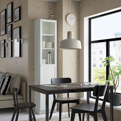 Comedor moderno con estantería blanca BILLY, mesa, sillas y sofá negros, paredes de madera, lámpara colgante, reloj y planta.