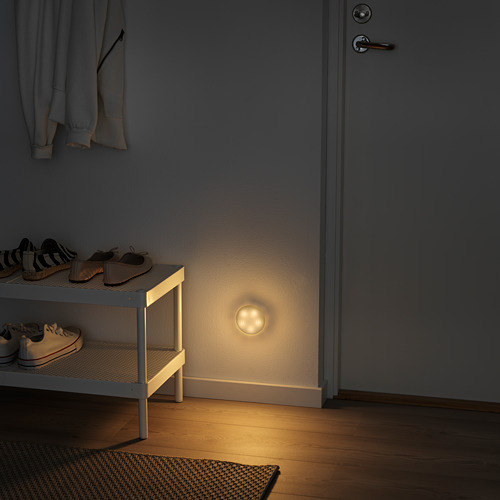 Pasillo oscuro con luz MOLGAN montada en la pared, zapatero y chaquetas colgantes. La luz es pequeña, redonda y blanca, iluminando el suelo.