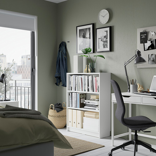Un dormitorio sereno con una estantería blanca BILLY que almacena libros y decoración, colocada entre una cama y un escritorio con una silla de oficina negra.