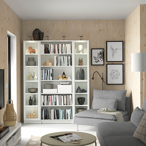 Sala de estar moderna con estantería BILLY blanca, sofá gris, mesa de centro y paredes de madera. La decoración incluye libros, jarrones y obras de arte enmarcadas.