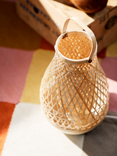 Una linterna de bambú tejida, llamada MISTERHULT, se encuentra sobre una superficie colorida. Su mango circular y su patrón de rombos crean un ambiente cálido y acogedor.