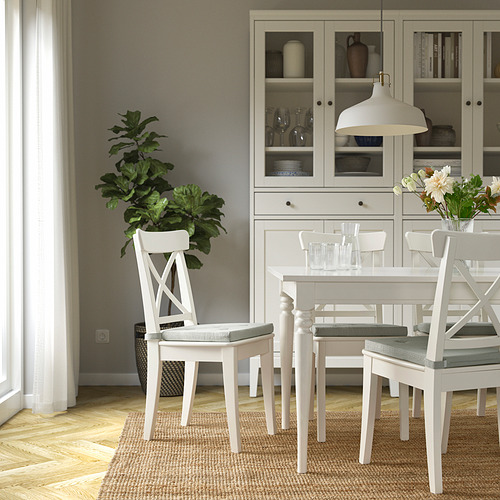 Mesa de comedor blanca con sillas JUSTINA, acolchada. Conjunto con vasos y flores. Habitación minimalista y acogedora con piso de madera, alfombra, gabinete y planta.
