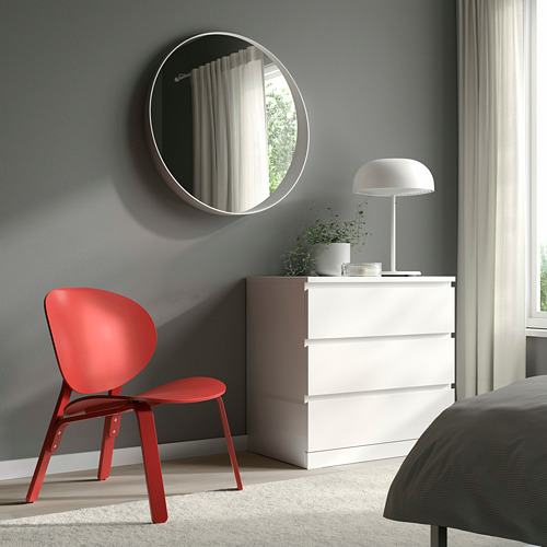 Dormitorio moderno con silla FRÖSET roja, tocador blanco, planta y espejo redondo. Paredes de color gris claro. Cama parcialmente visible. Uso eficiente del espacio.