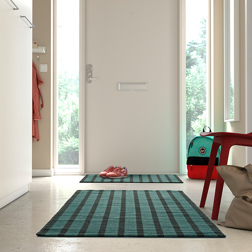 Entrada moderna con una gran alfombra a cuadros azul verdoso, una colchoneta más pequeña a juego, una silla roja para niños y una mochila verde cerca de puertas blancas.