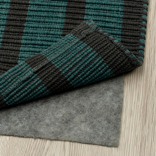 Una alfombra de rayas verdes y negras oscuras, colocada sobre una almohadilla gris sobre un suelo de madera. La alfombra tiene una superficie plana, lo que facilita la aspiración.