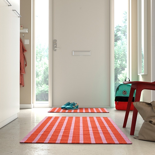 Alfombra de rayas rosas y naranjas brillantes cerca de la puerta, más pequeña que la silla y la bolsa.