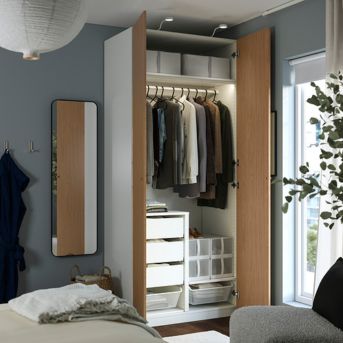Un moderno armario de madera llamado PAX se encuentra abierto, mostrando espacios de almacenamiento internos que incluyen perchas con ropa, cajones y estantes con cestas.