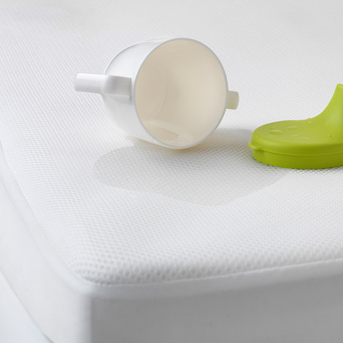 Taza blanca volcada sobre un protector de colchón blanco con un vasito verde cerca.