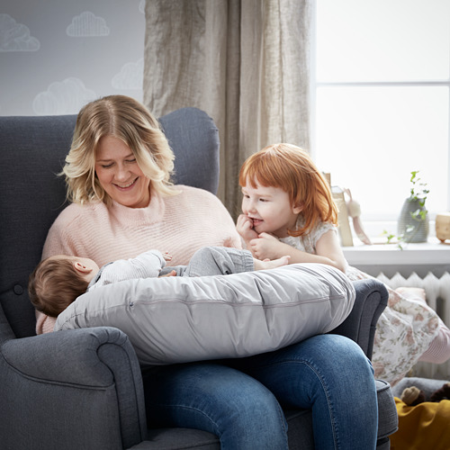 Una mujer y dos niños disfrutando de un momento acogedor en un sofá gris con una almohada de lactancia, rodeados de una decoración suave en una habitación bien iluminada.
