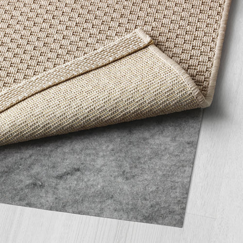 Una alfombra tejida de color beige con una esquina doblada, colocada sobre una base de alfombra de fieltro gris, adecuada para uso en interiores y exteriores.