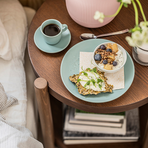 La mesa de madera sostiene un plato de color menta con comida y una taza de café. Una cama con almohadas está al lado.