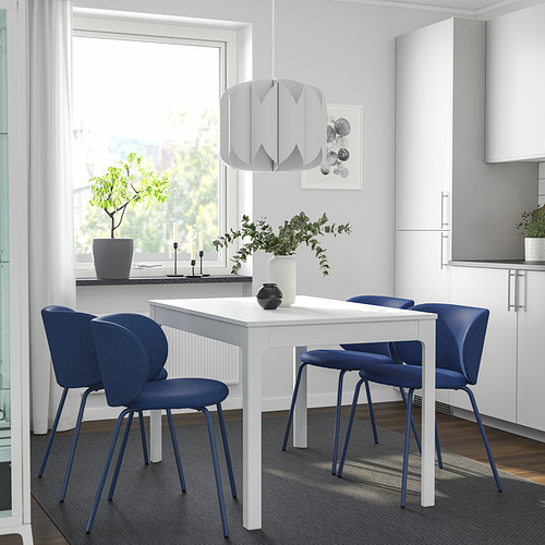 Comedor moderno con mesa blanca, sillas azules, gran ventanal, planta y lámpara geométrica.