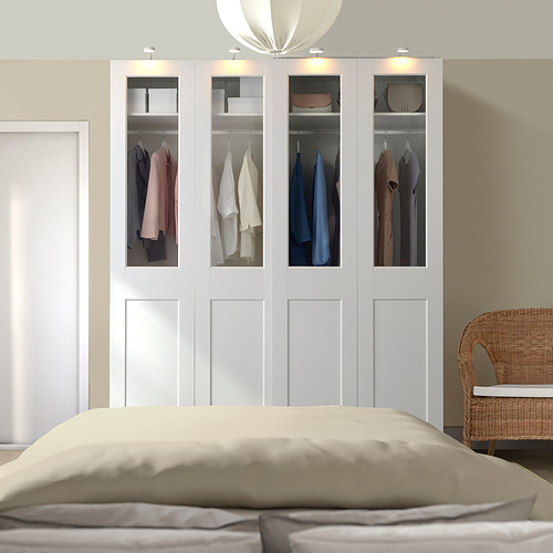 Armario PAX blanco con puertas de cristal, exhibiendo ropa. Alto, moderno y eficiente, junto a cama y silla de mimbre.