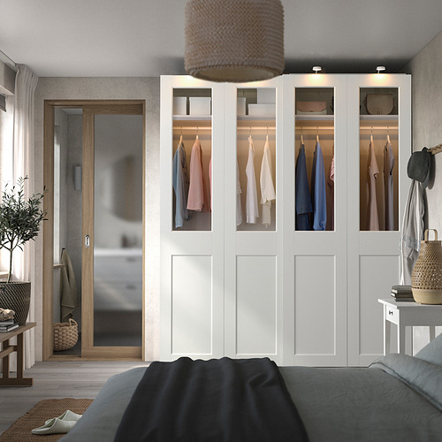 Dormitorio moderno con gran armario blanco PAX, puertas transparentes, ropa tendida, junto a la puerta de madera, cama negra, cesta de mimbre y planta.