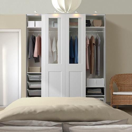 Un gran armario blanco PAX con cristal, que exhibe ropa y artículos organizados. Se alza junto a una silla de mimbre y una cama con ropa de cama beige.