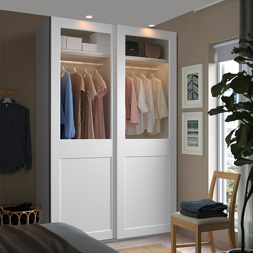 El armario moderno con puertas de vidrio muestra la ropa, mezclándose con las paredes beige y los grandes ventanales. A su lado, una silla sostiene ropa doblada.