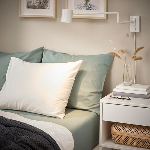 Acogedor rincón de dormitorio con almohada DVALA gris verdosa, sábanas azul claro, almohadas blancas, libros, flores secas y lámpara de pared.