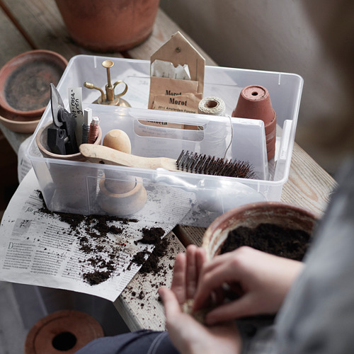 Caja de almacenamiento de plástico transparente con compartimentos, que contiene herramientas de jardinería y macetas, colocada sobre papel de periódico con tierra.