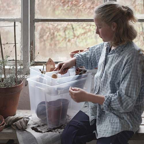 La mujer está sentada junto a la ventana, manipulando artesanías en una caja de plástico transparente SAMLA. Lleva una camisa a cuadros, centrándose en las miniaturas. Planta en maceta y guantes cerca.