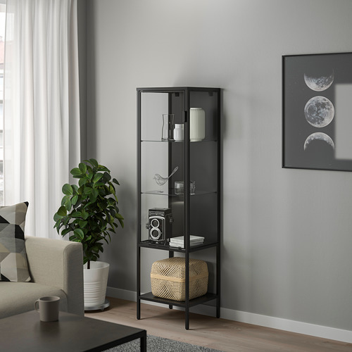 Mueble con puerta de cristal RUDSTA negro en un salón moderno, con cámara vintage, junto a grandes plantas y arte de las fases lunares.
