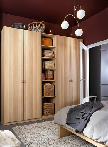 El armario PAX de madera natural se encuentra al lado de la cama. Las puertas abiertas muestran cestas de mimbre en los estantes. La habitación tiene paredes de color burdeos, luces blancas.