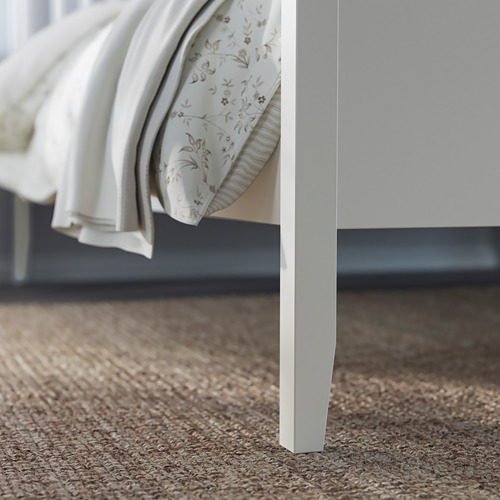Una esquina de un marco de cama IDANÄS blanco sobre una alfombra beige. La cama tiene un diseño elegante y moderno con una cabecera robusta y minimalista.