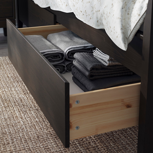 Esta imagen muestra un marco de cama marrón oscuro de IDANÄS con cajones de almacenamiento de madera, mantas grises cuidadosamente dobladas en el interior.