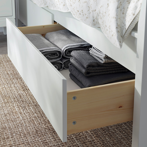 Un cajón de madera blanca debajo de una cama sostiene cuidadosamente toallas grises dobladas.