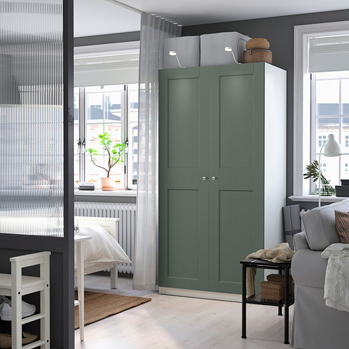 Esta imagen es un armario PAX verde en una habitación moderna. Se alza alto, junto a un sofá y una estantería, y ofrece un amplio espacio de almacenamiento con sus dos puertas.
