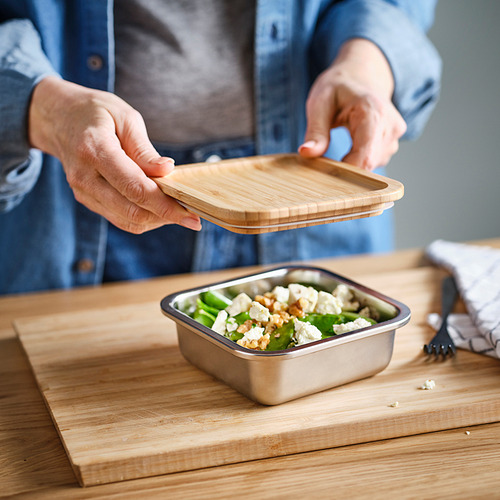 Persona colocando una tapa de bambú en un recipiente de acero inoxidable lleno de ensalada sobre una tabla de cortar de madera.