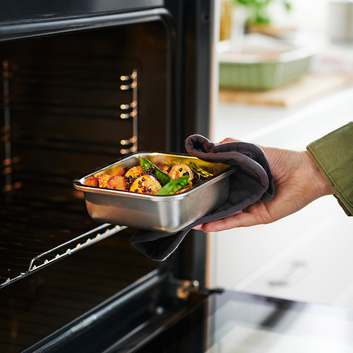 Una mano coloca un recipiente metálico de comida con verduras en un horno. El recipiente se utiliza para almacenar las sobras y recalentar los alimentos.