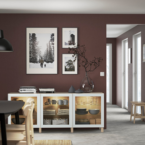 White BESTÅ sideboard with glass doors, displaying dishes and vases, under three framed artworks on a mocha wall.
