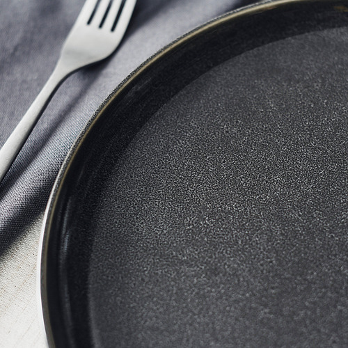 Plato de gres GLADELIG negro con esmalte texturizado junto a un tenedor sobre un paño oscuro.
