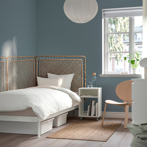 Dormitorio con cabecero de ratán azul TOLKNING, ropa de cama blanca, silla de madera, mesa auxiliar con libros y ventana.