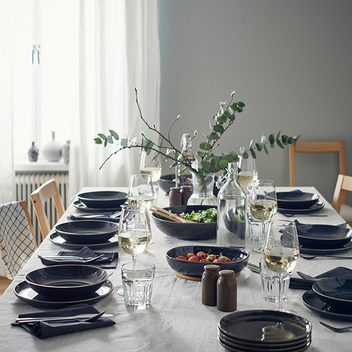 Elegante configuración de comedor con platos profundos de color gris oscuro GLADELIG, vasos transparentes, servilletas cuidadosamente dobladas y un jarrón central con vegetación.