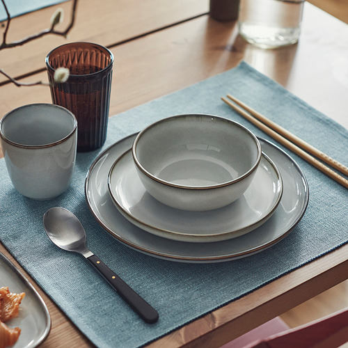 Mesa con placa auxiliar, cuenco, taza, palillos y cuchara de gres gris GLADELIG sobre estera azul.